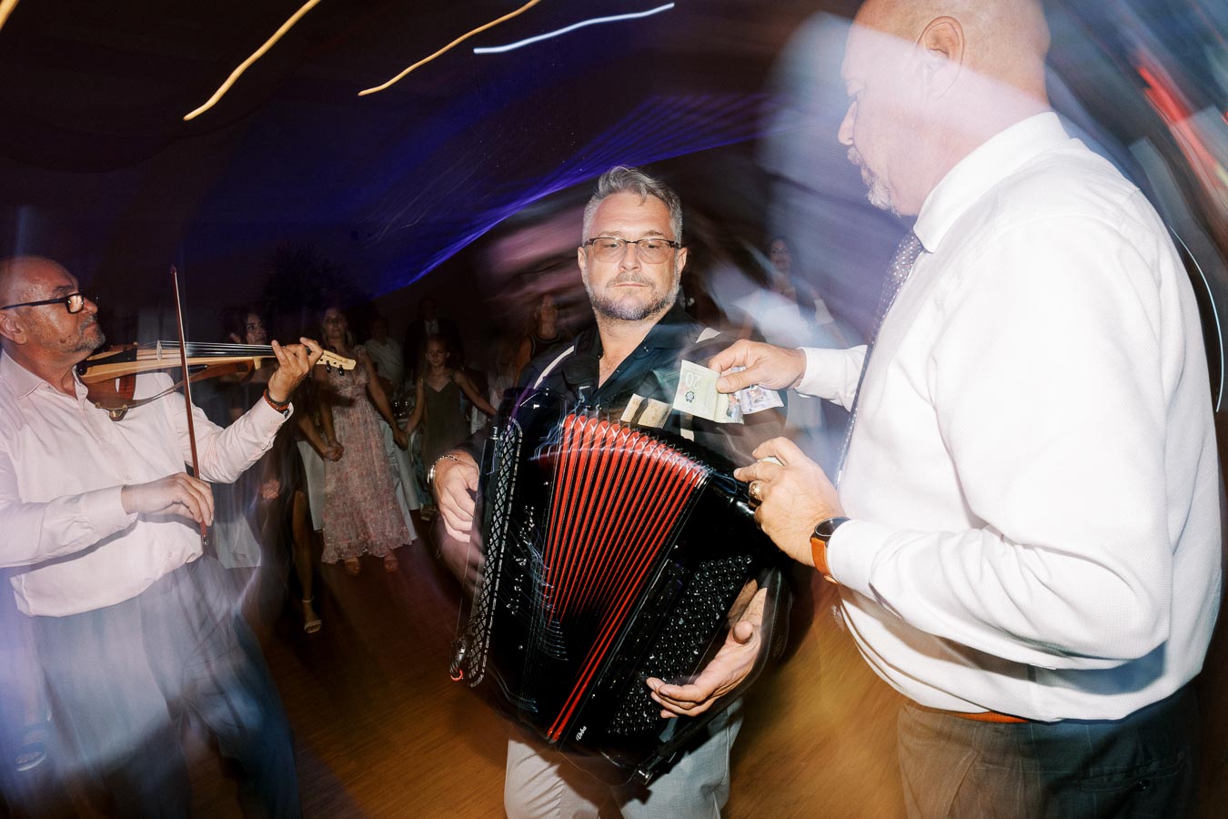 A man playing an accordion at a lively event, with another man handing him money, while a violinist performs nearby. The blurred background suggests movement and dancing guests, emphasizing the festive atmosphere.