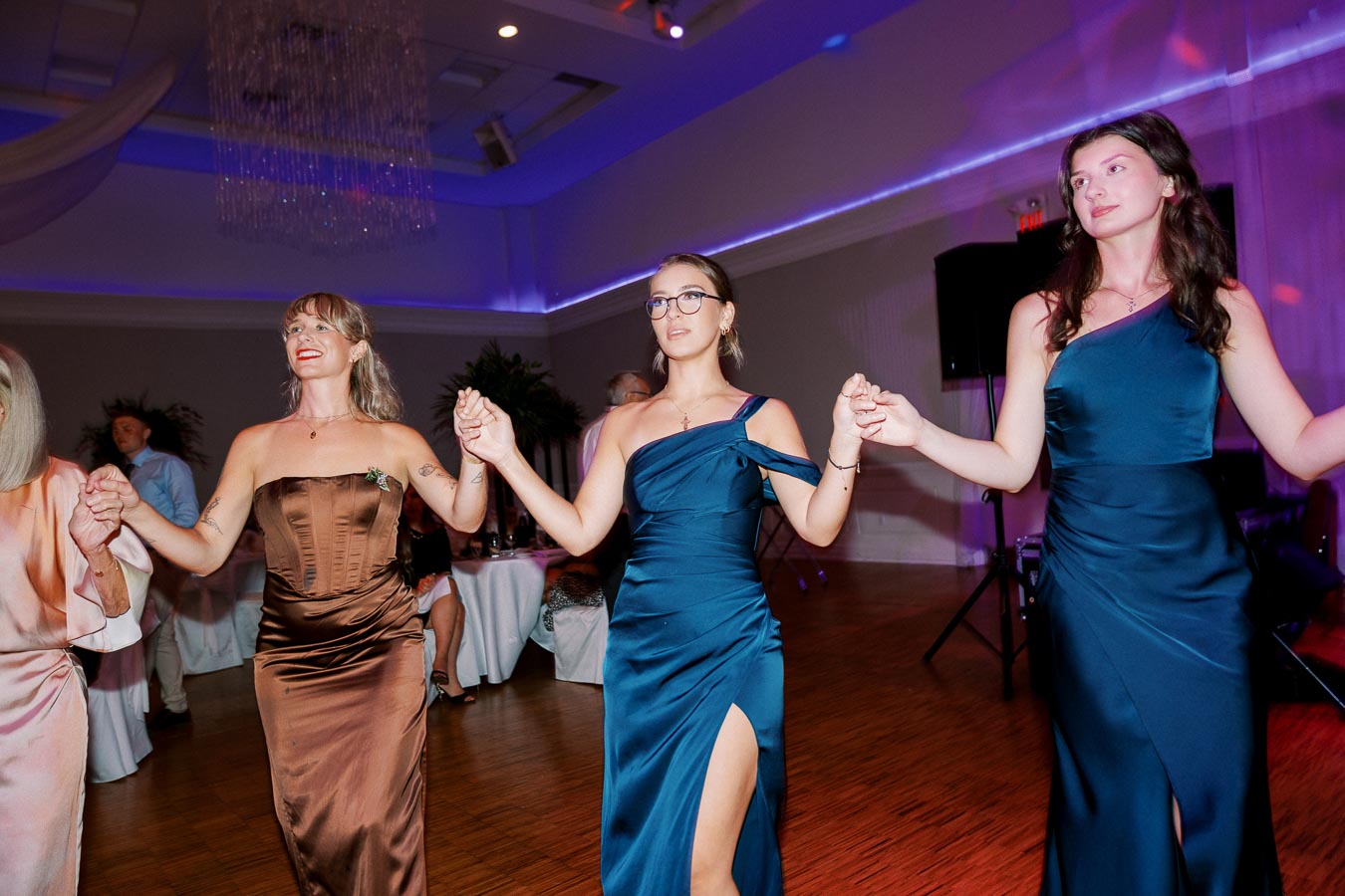 Three women in elegant gowns dancing at an indoor event, holding hands and smiling, with a decorated venue in the background.