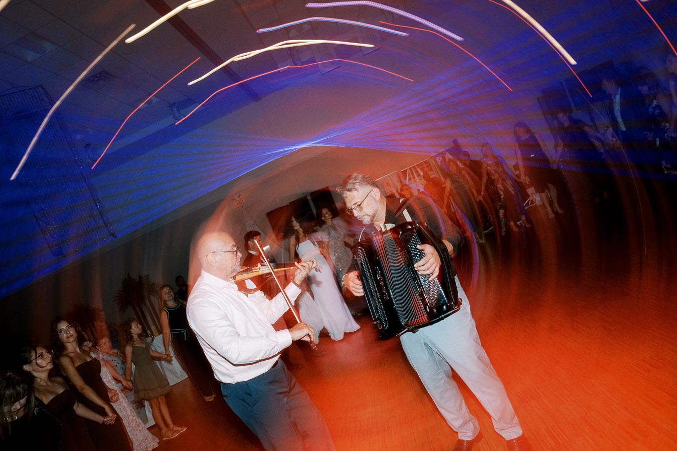 Blurred motion photo of two musicians, one playing a violin and the other an accordion, at an event with dancing guests and vibrant lighting effects.