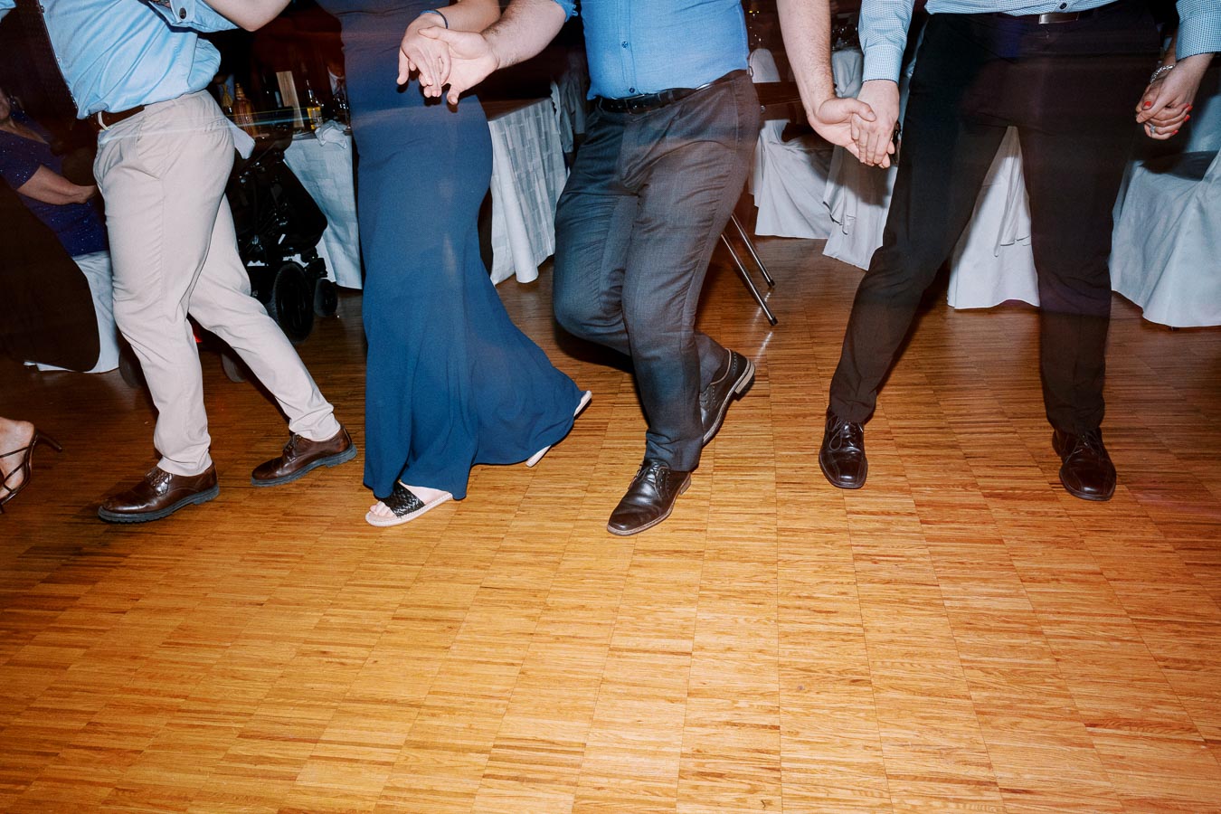 Four people in formal attire holding hands and dancing in a line on a wooden floor during a lively event.