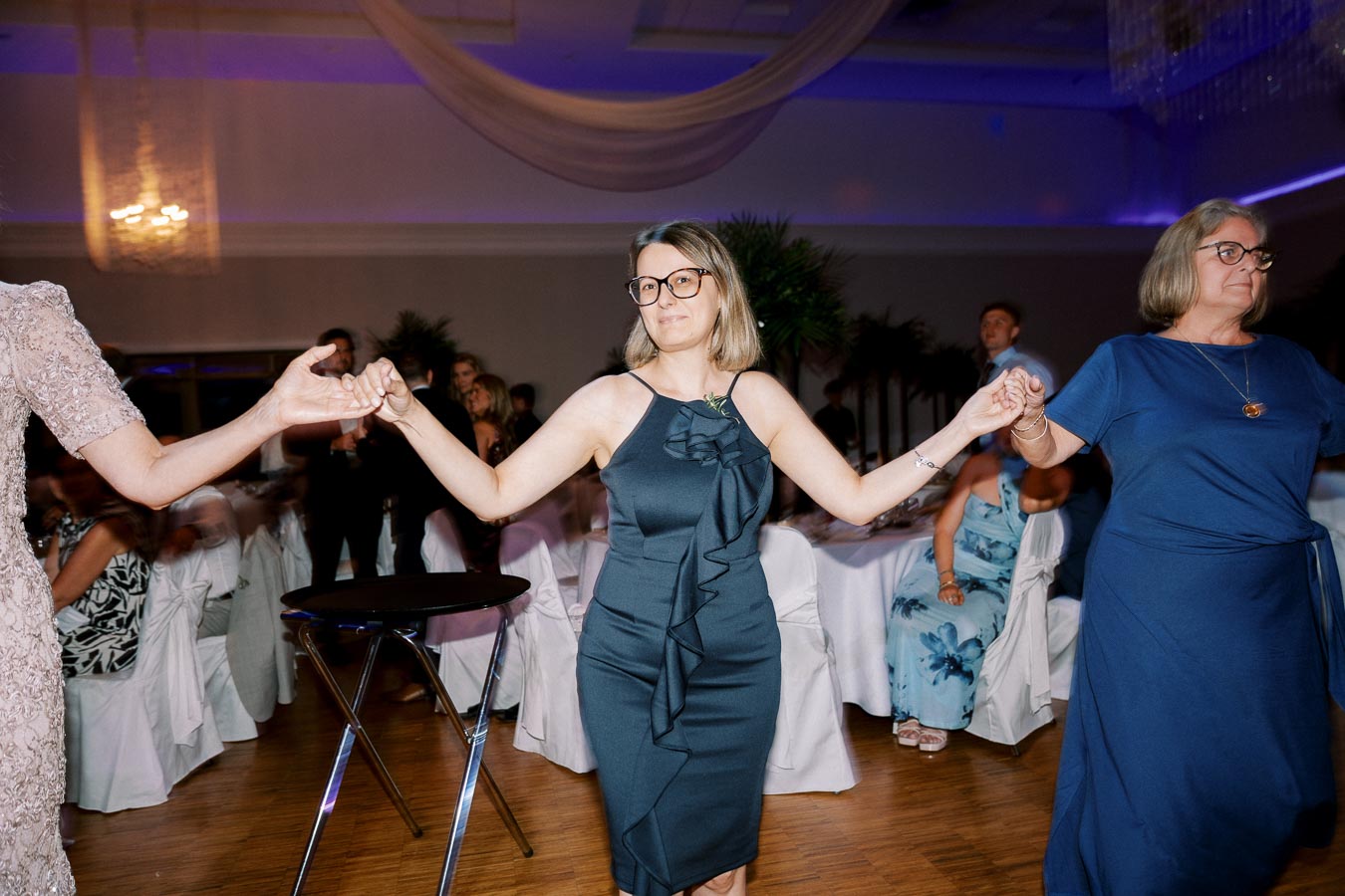 A group of people holding hands and dancing in a well-decorated banquet hall with elegant lighting and dressed-up attendees, capturing a joyful moment at a formal event or celebration.