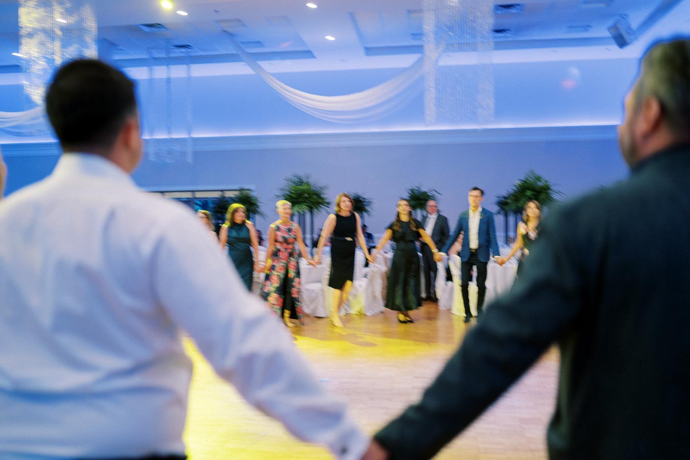 A group of people hold hands in a circle on a dance floor at an elegant, blue-lit event with chandeliers and potted plants.