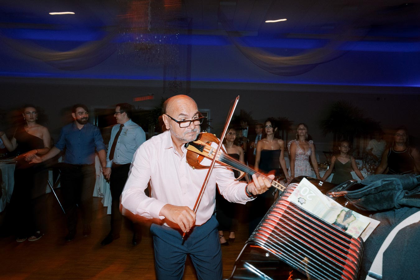 A lively event scene with a man passionately playing the violin, engaging the audience on the dance floor at a wedding reception. The room is dimly lit with blue ambient lighting, highlighting the festive atmosphere.