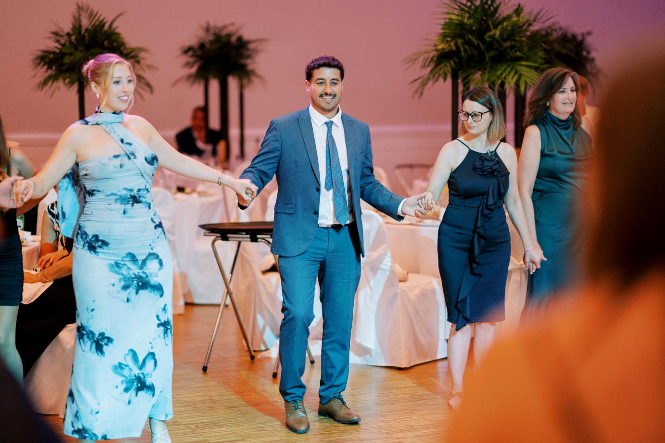 A group of people holding hands in a circle at a formal event, smiling and wearing elegant attire, with decorative plants and a wooden floor in the background.