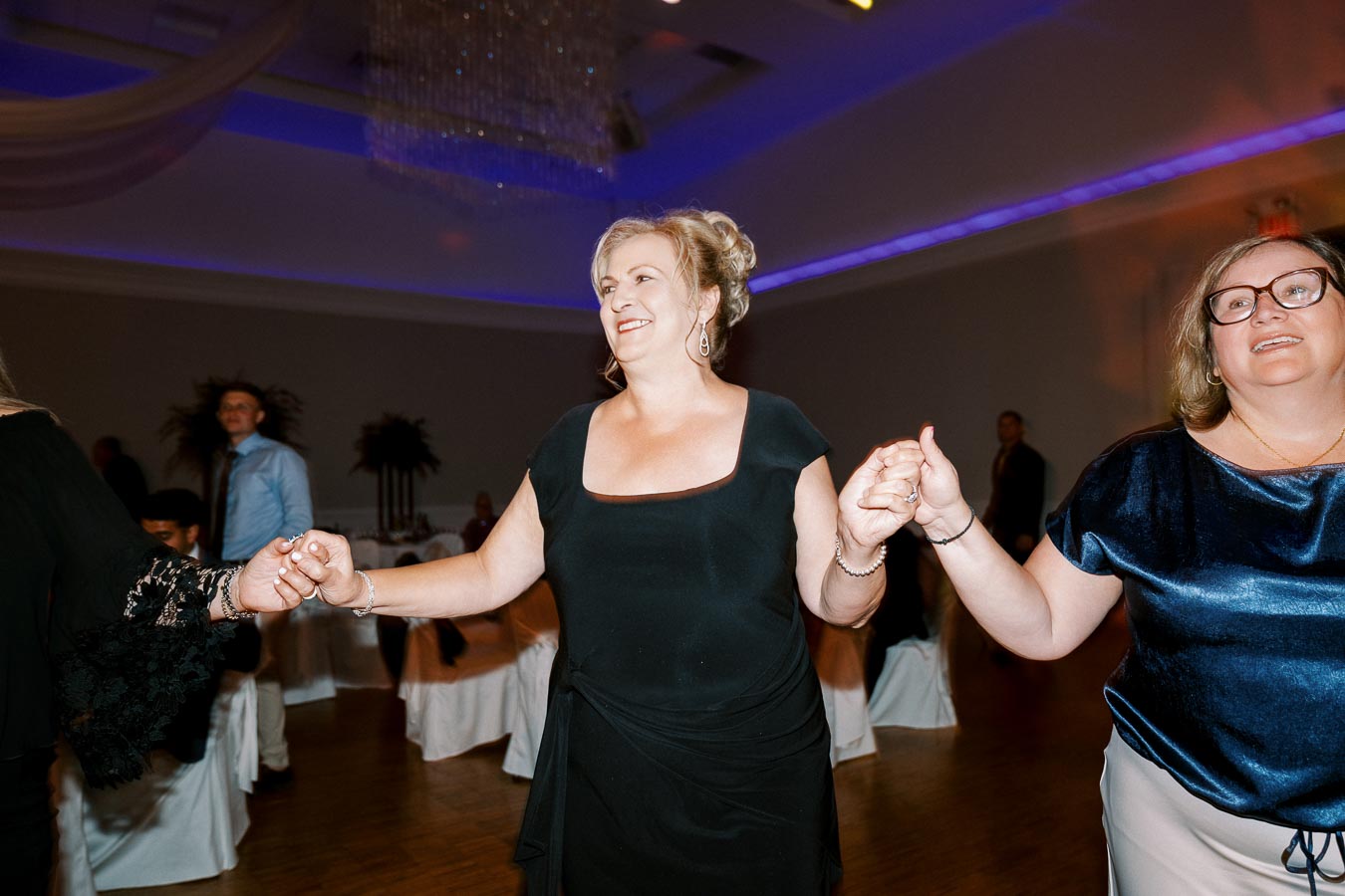 A group of people holding hands and smiling while dancing at a lively indoor event, with elegant lighting and decor in the background.