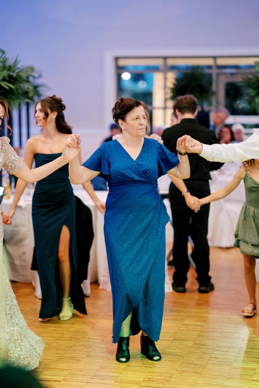 A group of people dancing in a circle at a celebration, with a woman in a blue dress holding hands and smiling, surrounded by attendees in formal attire.