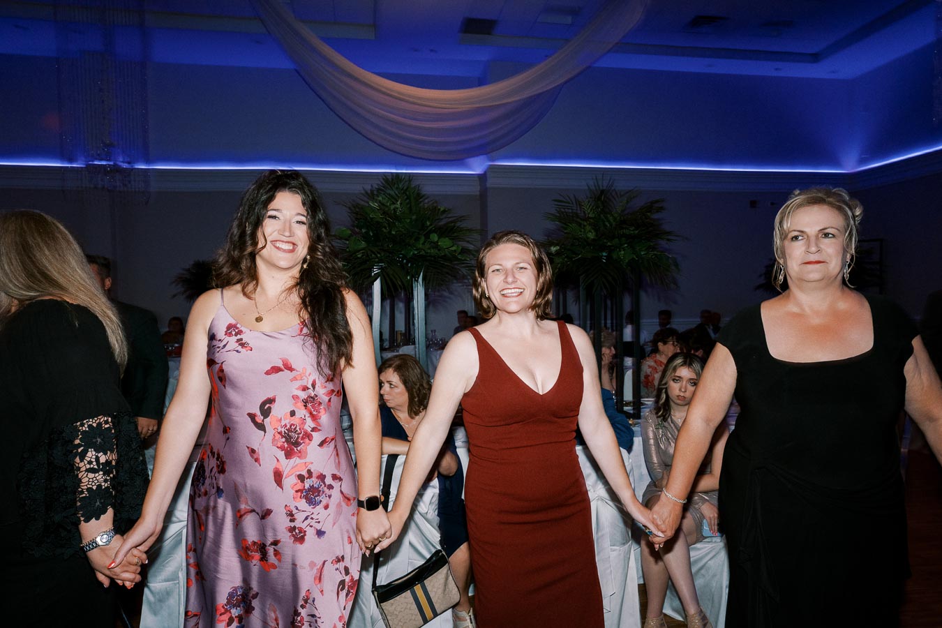 Three women in elegant dresses holding hands and smiling at a formal event, with a blue-lit room and seated guests in the background.