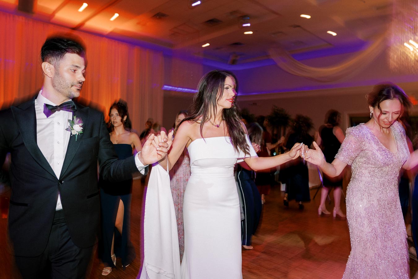A group of elegantly dressed people dancing at a wedding reception, with a man in a tuxedo and women in formal dresses, celebrating joyfully under colorful lighting.