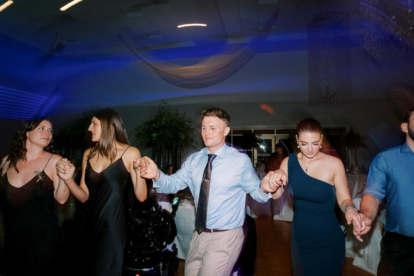 People dancing in a circle at a party or wedding reception, holding hands, wearing formal attire.