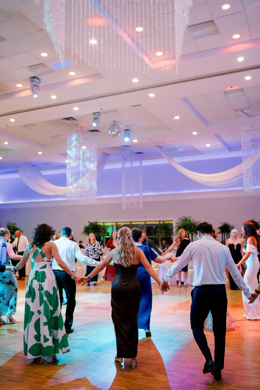 A group of elegantly dressed people dancing in a circle at a brightly lit indoor wedding reception, featuring chandeliers, draped fabric, and vibrant lighting on a spacious wooden dance floor.