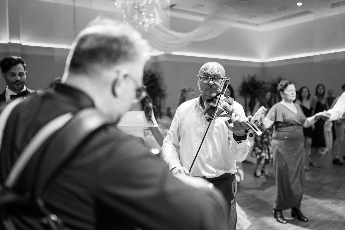 Black and white photo of a lively wedding reception where a musician in a white shirt plays the violin among a group of dancing guests, creating an engaging and joyful atmosphere.