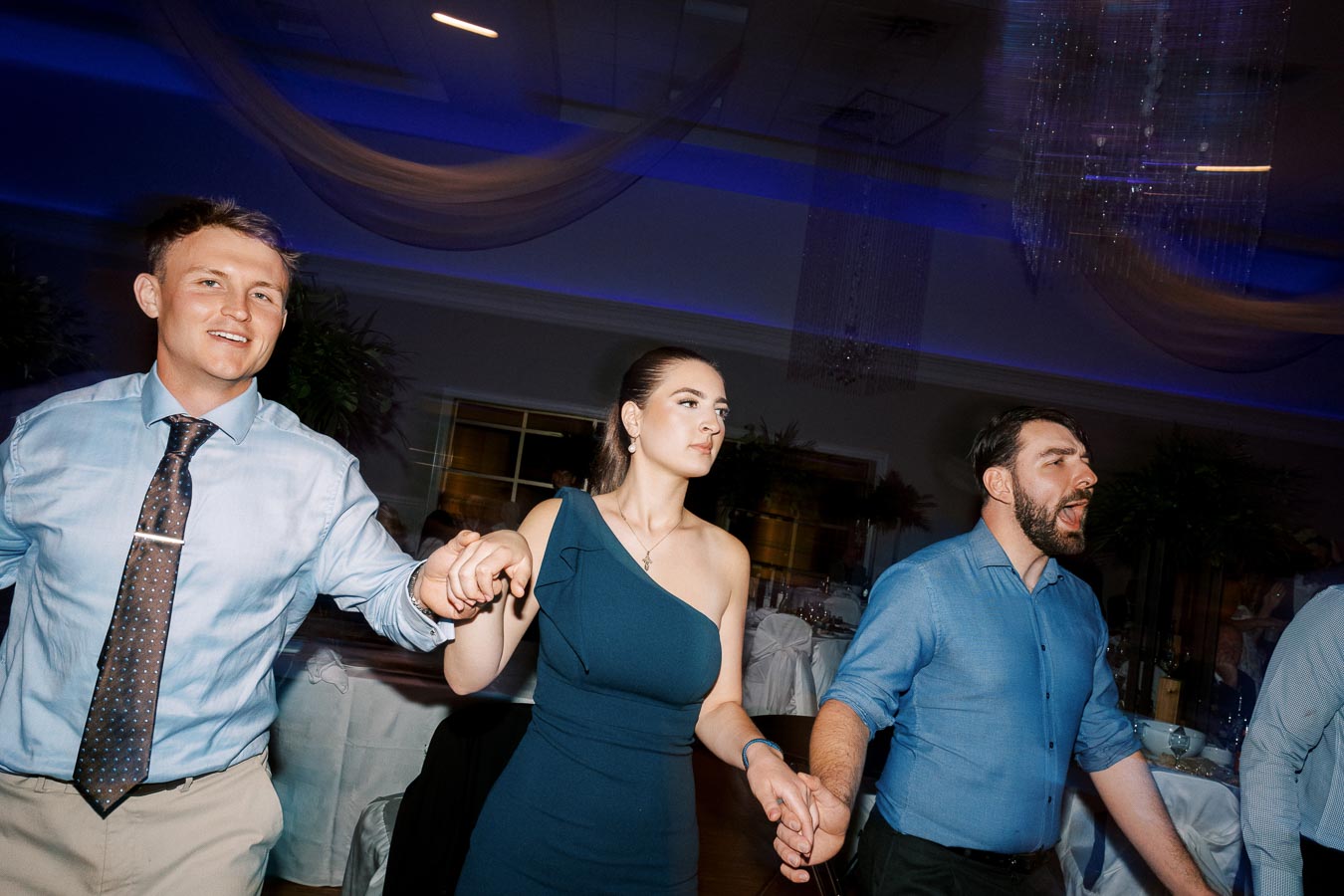 People enjoying a dance at a party, illuminated by blue lighting, wearing formal attire and holding hands as they move in unison.