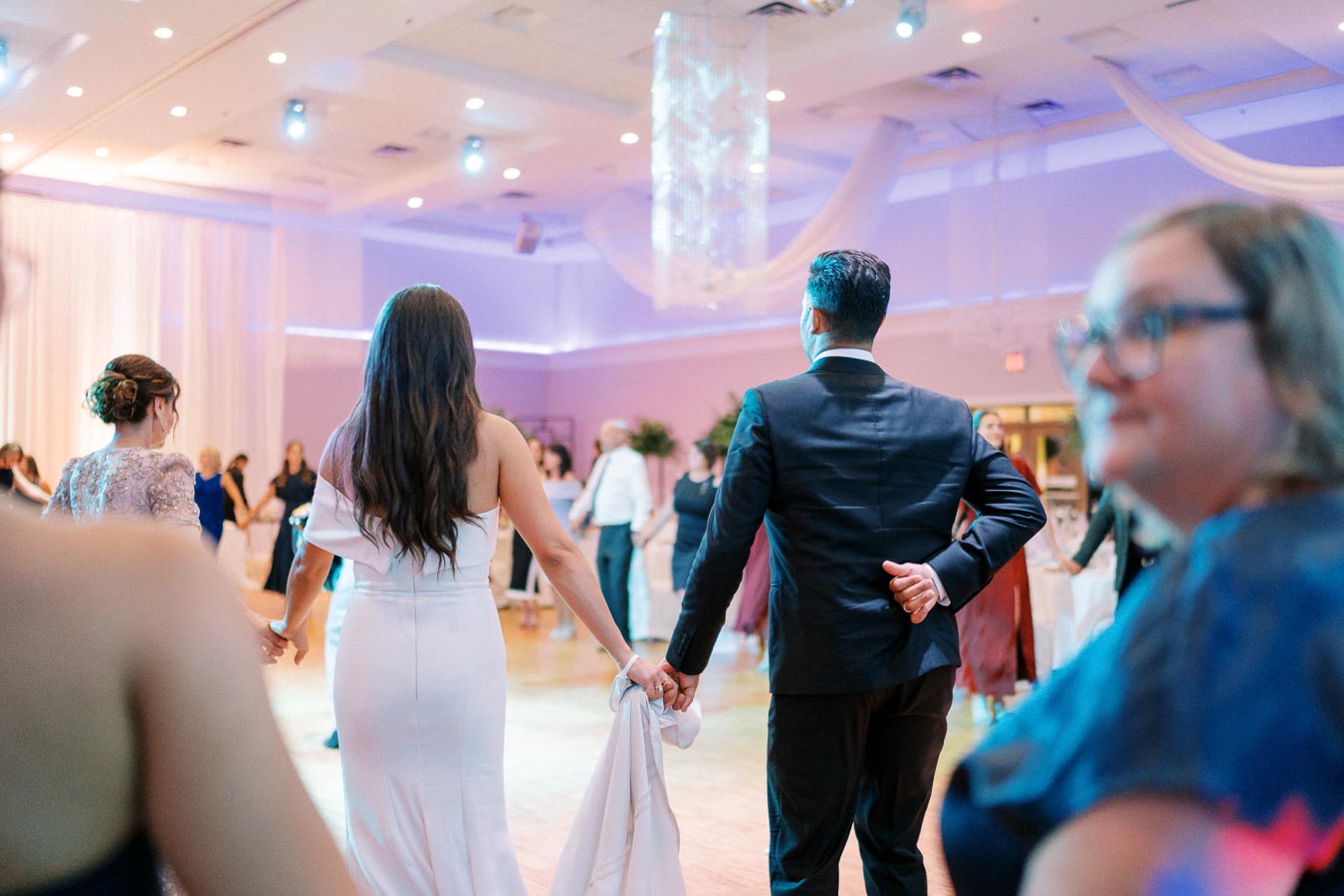 A couple dancing at a wedding reception, holding hands on a lively dance floor, with guests enjoying the celebration in a beautifully lit event space.