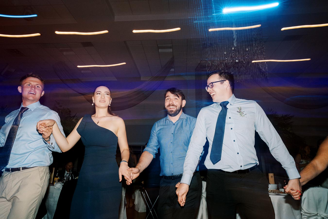 A group of people enjoying a lively dance at an indoor event, holding hands and smiling, with colorful blurred lights in the background adding to a festive atmosphere.