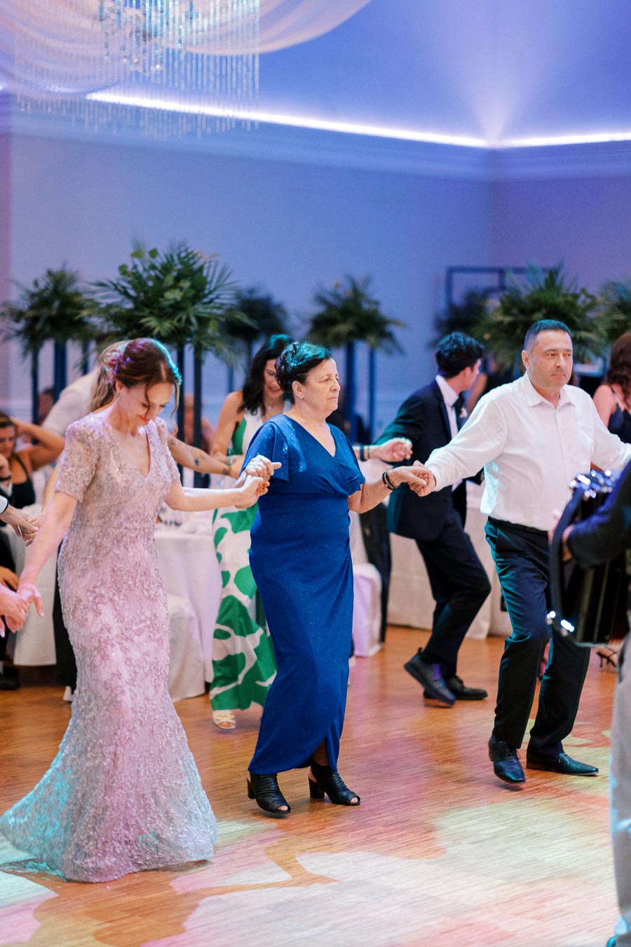 A group of elegantly dressed people dancing at a formal event, illuminated by ambient lighting and surrounded by decorative plants.