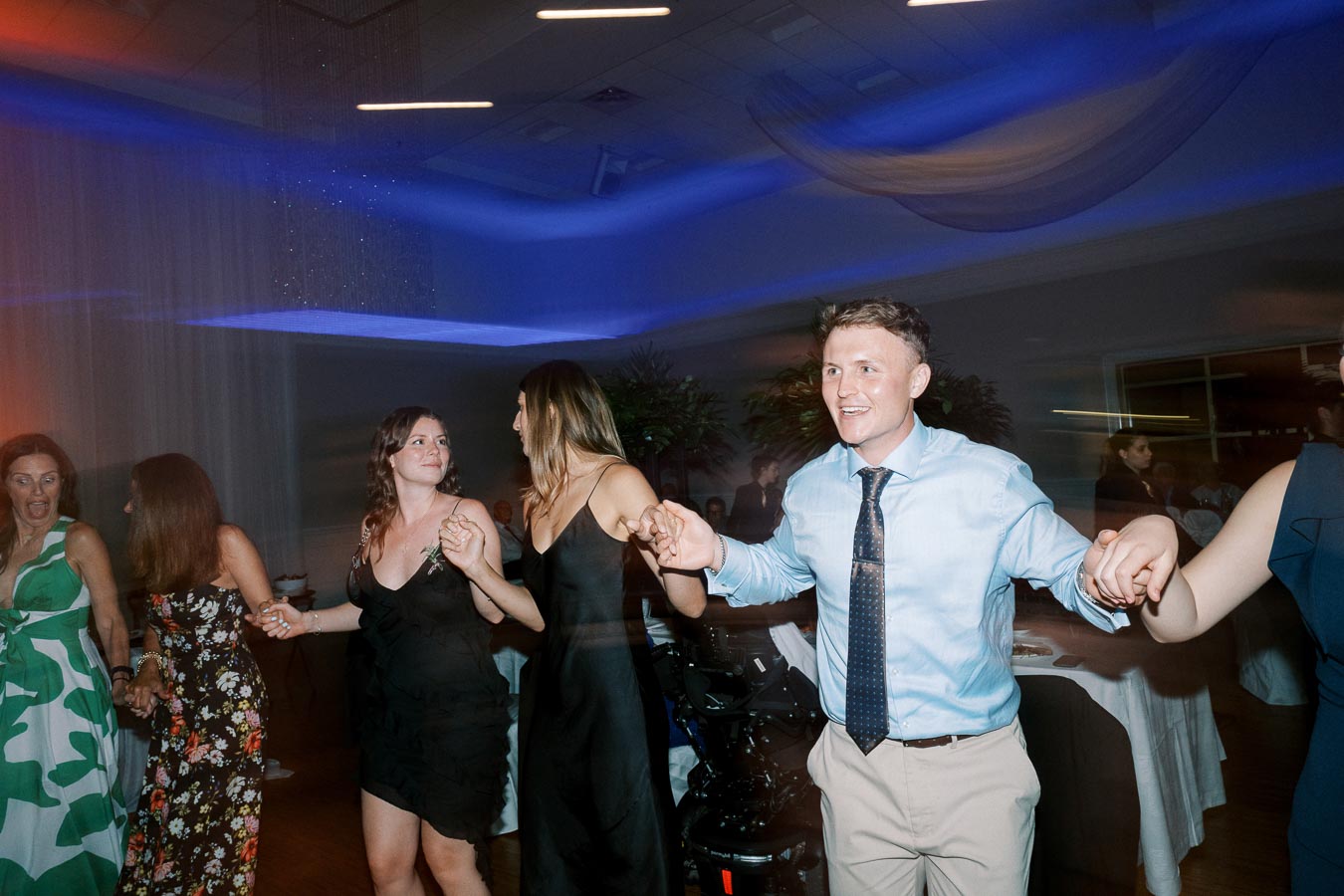 Group of people joyfully dancing hand in hand at a festive event, illuminated by colorful lights in a lively indoor setting.