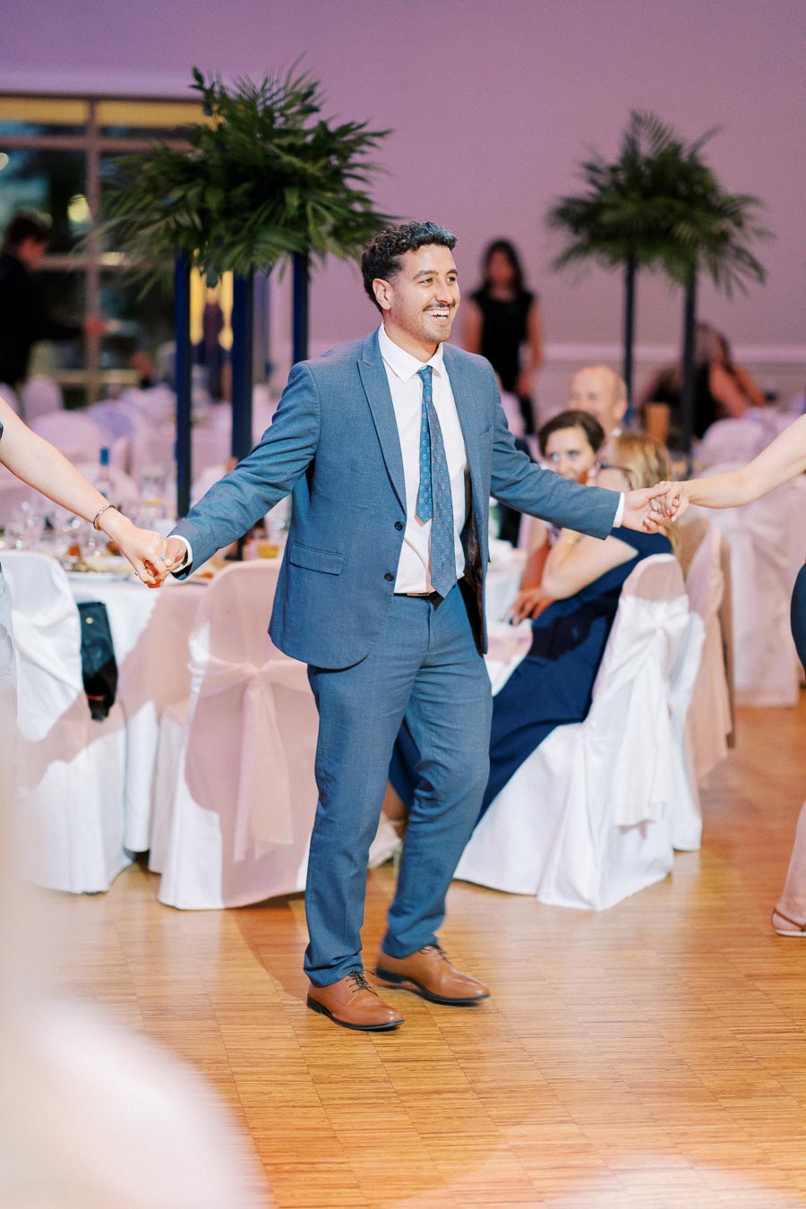 A man in a blue suit joyfully dancing at a formal event, holding hands with partners on either side, with elegantly decorated tables and green plants in the background.