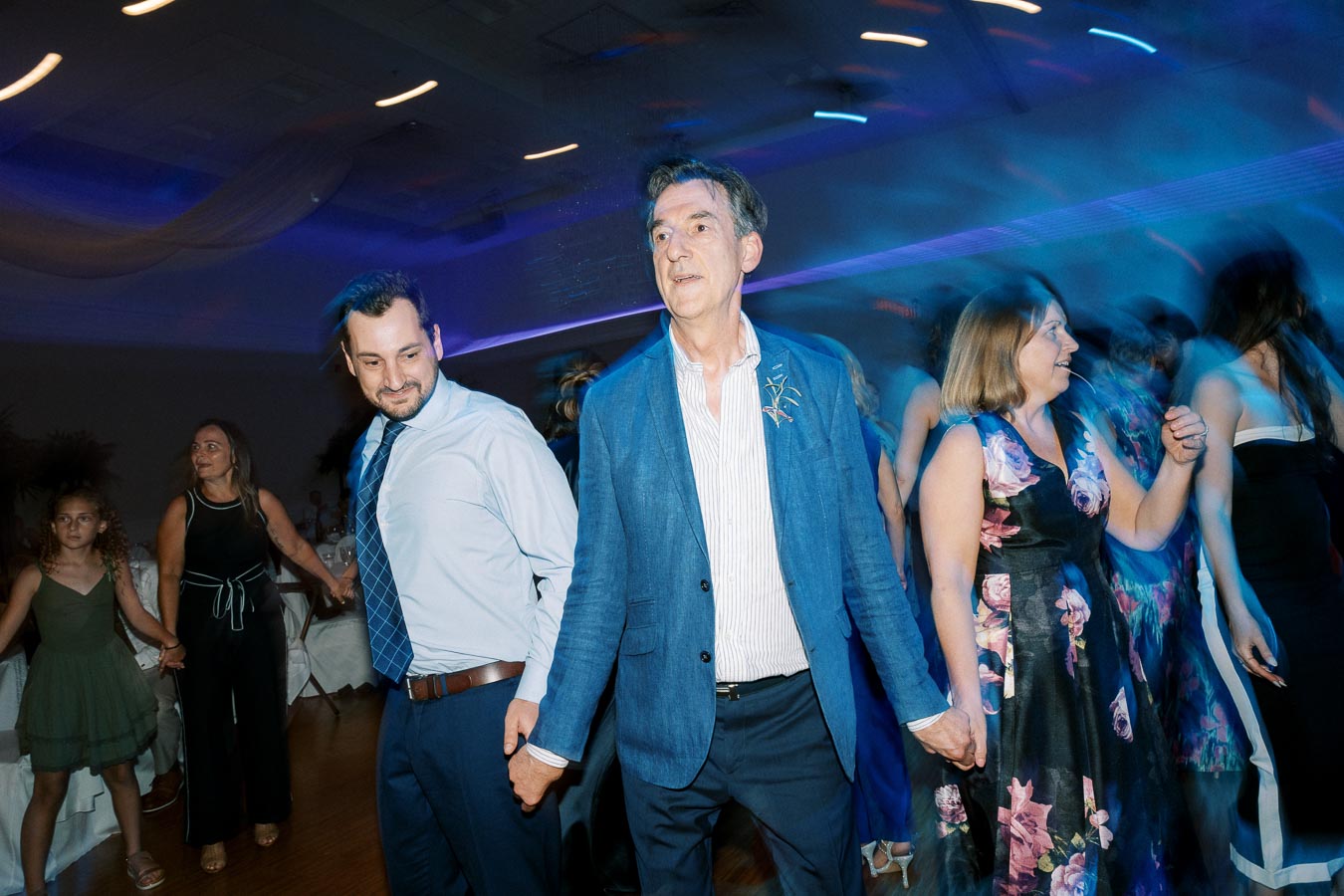 A group of people enjoying a lively dance at a wedding reception, dressed in formal attire, under colorful lights.