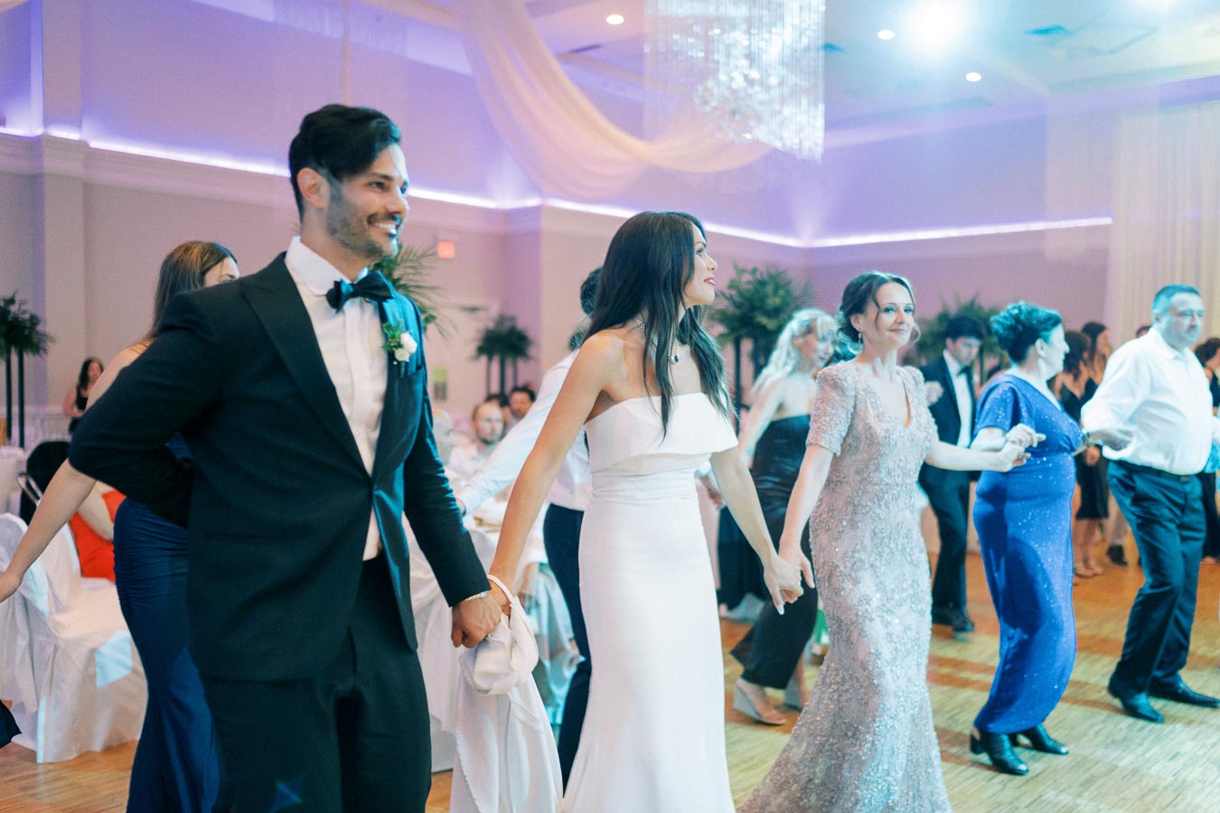 Group of elegantly dressed people dancing and holding hands at a wedding reception, featuring a man in a tuxedo and women in stylish gowns, in a beautifully decorated hall with ambient lighting.