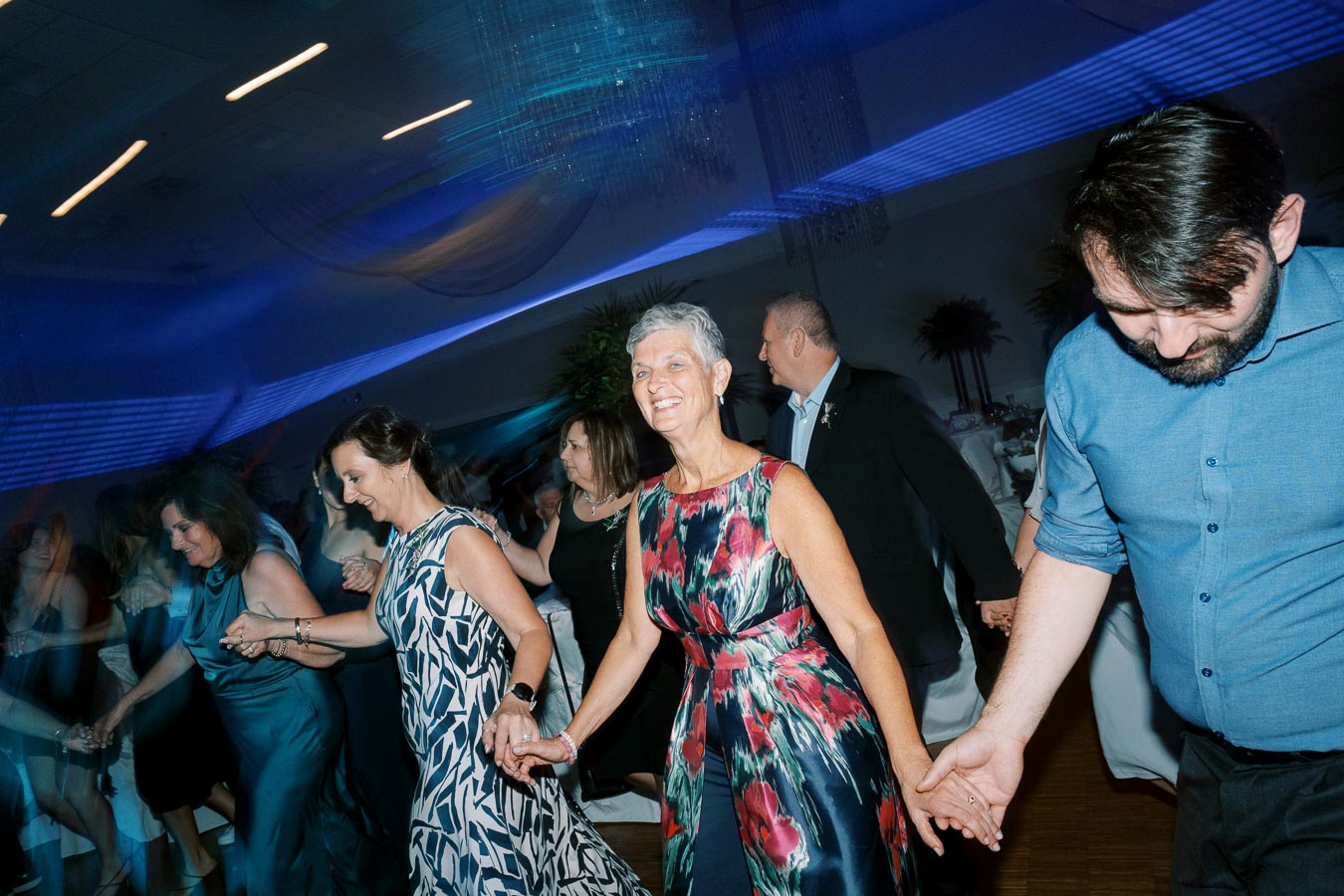 People dancing joyfully in a ballroom with vibrant lighting, wearing elegant dresses and formal attire, holding hands.