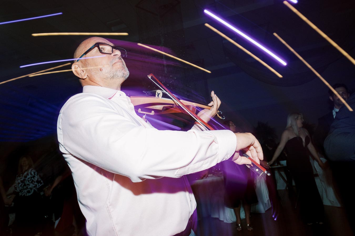 A man in a white shirt energetically playing a violin at a party, with streaks of colorful lights creating a dynamic atmosphere.