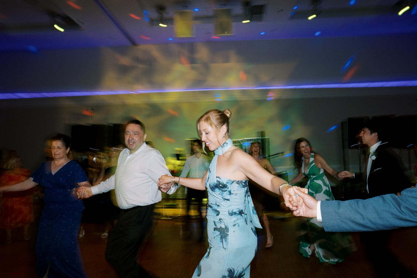 A festive wedding reception scene with joyful guests in formal attire dancing in a circle, surrounded by colorful lights creating a lively and celebratory atmosphere.