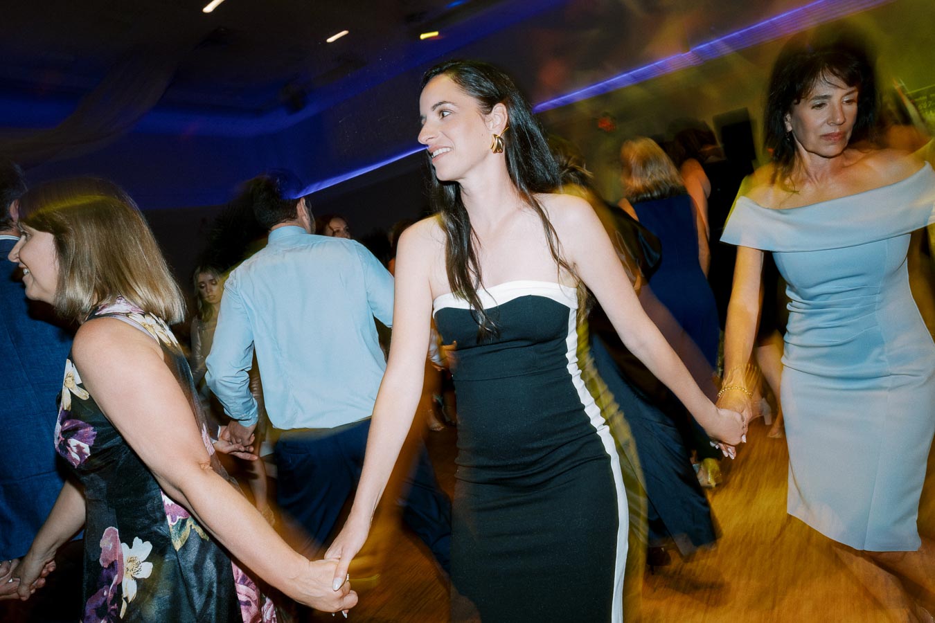 People joyfully dancing at a party, wearing elegant attire and holding hands, while colorful lights illuminate the room.