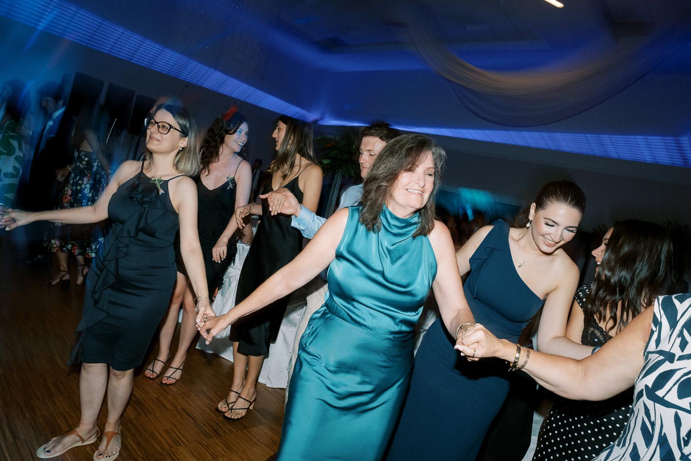 A group of joyful people dancing at a lively party, dressed in elegant evening attire, under vibrant blue lighting.
