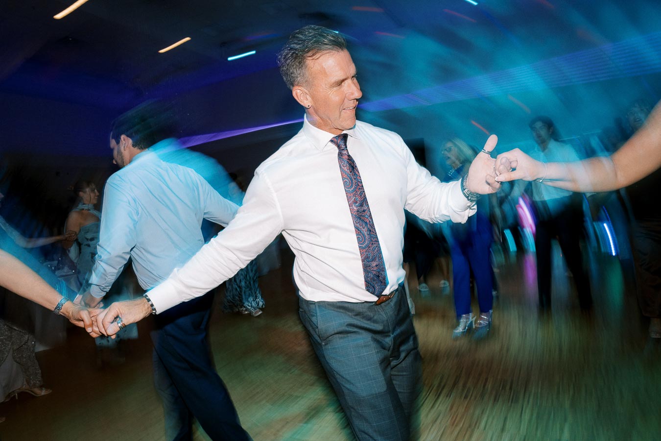 Man in formal attire dancing energetically at a party with colorful blurred lights in the background.