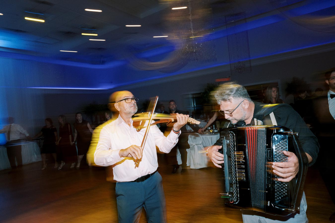A man playing the violin and another playing the accordion at a lively indoor event with blue ambient lighting and blurred motion effect, capturing the vibrant atmosphere.