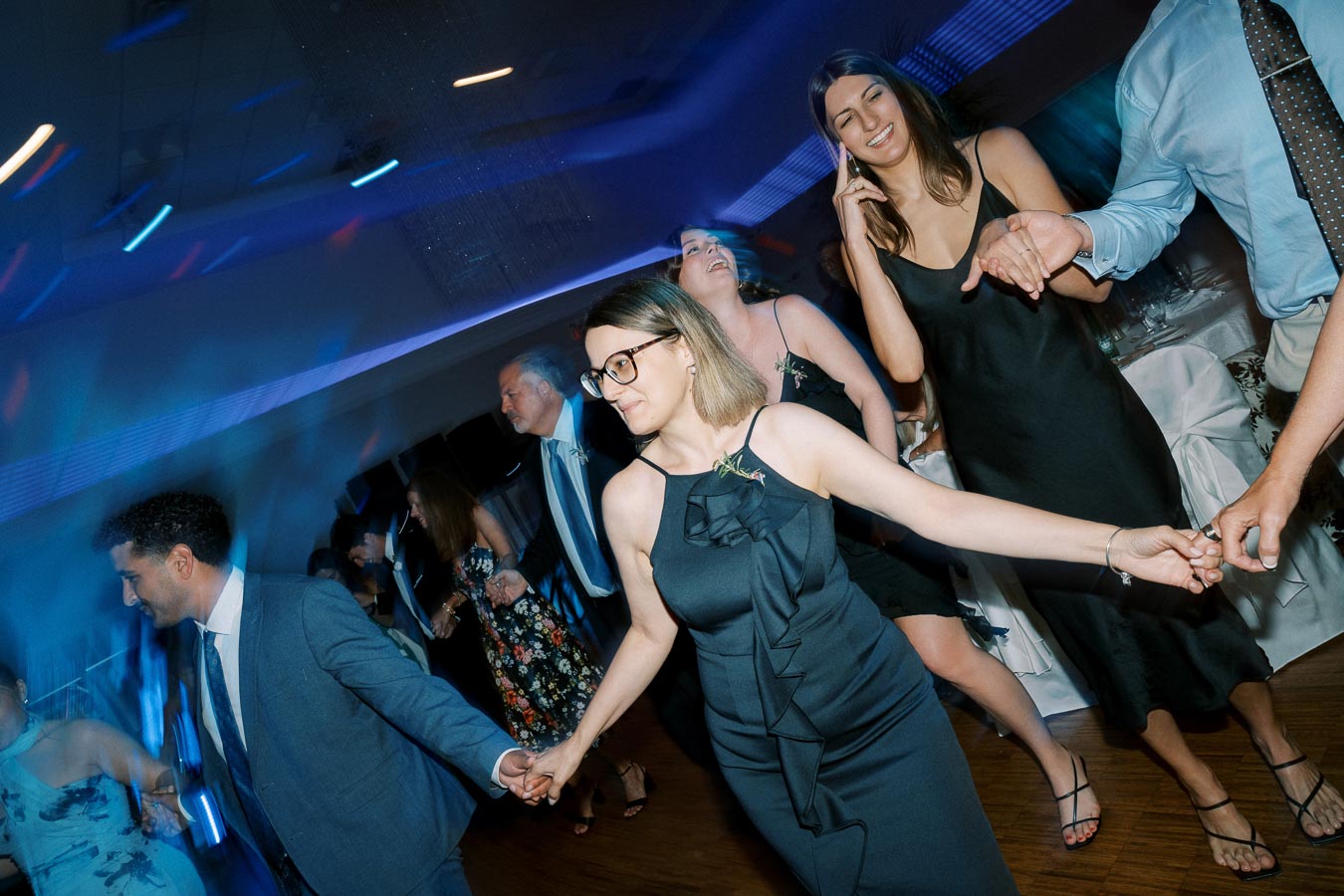 A group of people joyfully dancing in formal attire at an indoor event with vibrant blue and purple lighting, some holding hands and smiling.