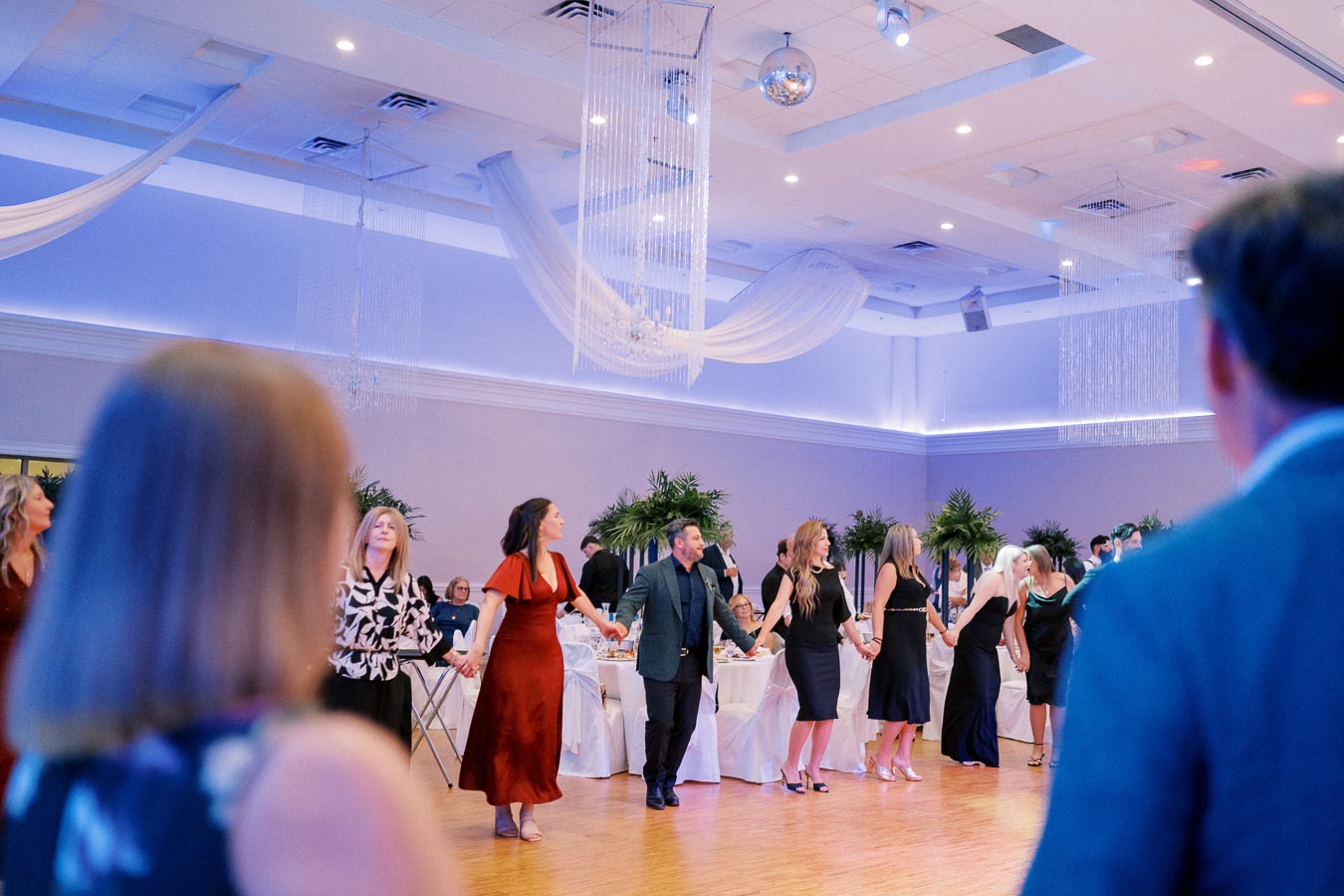 People dancing at a formal event in an elegantly decorated ballroom, featuring ambient blue lighting and hanging drapes, with banquet tables and greenery in the background.