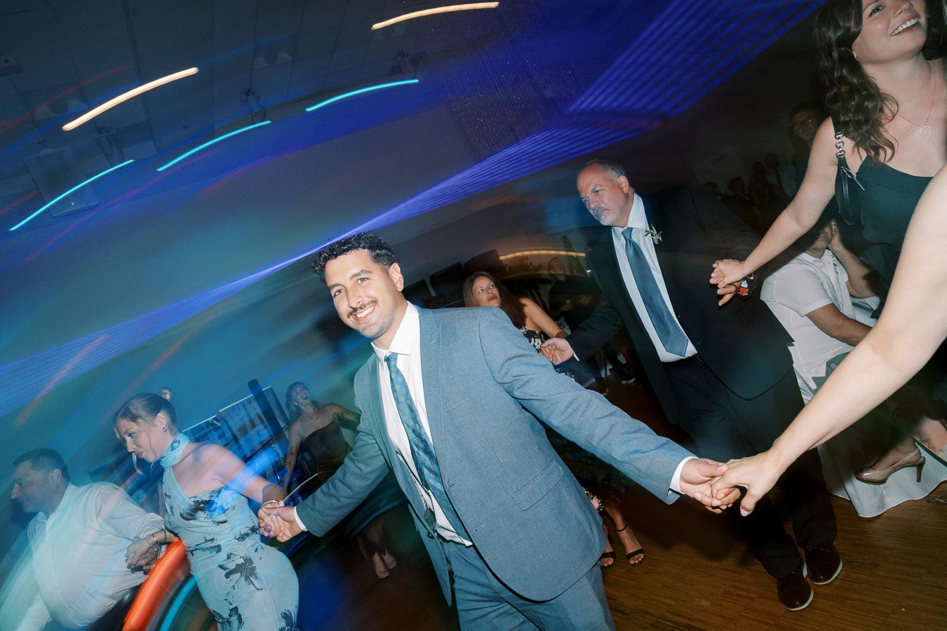 A joyful group of people dancing in a dimly lit room with colorful lights at a wedding reception, highlighting a festive and vibrant atmosphere.