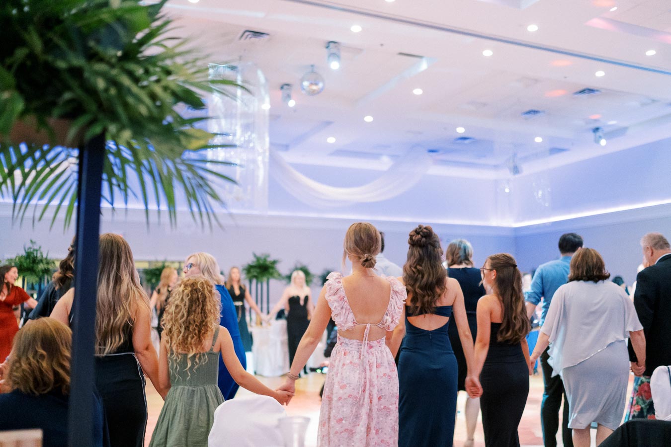 People gathered in a decorated venue, holding hands in a circle, with greenery and soft lighting creating an elegant atmosphere.