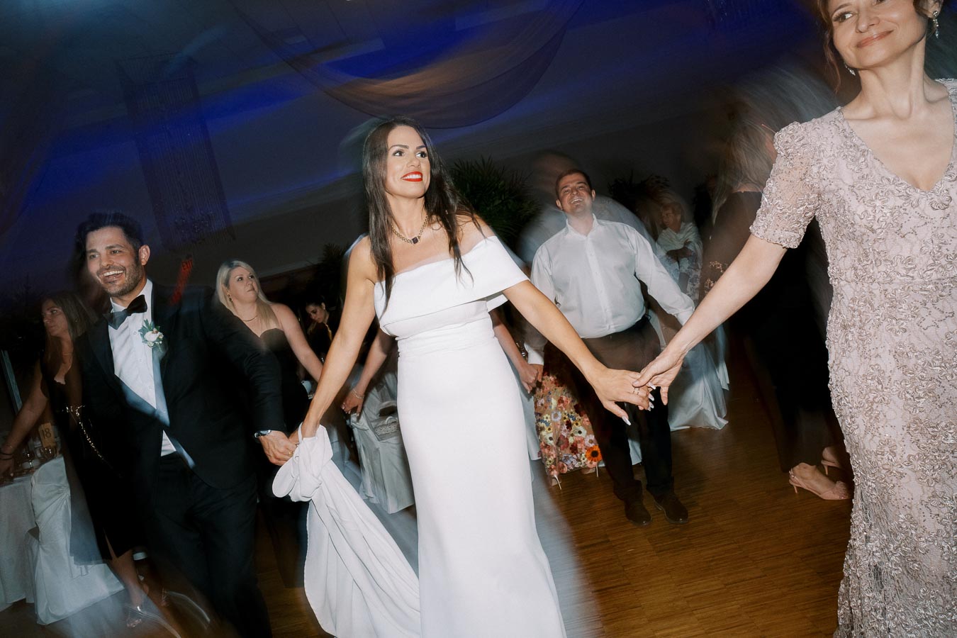 Wedding guests joyfully dancing at a reception, featuring a woman in an elegant off-the-shoulder white dress holding hands in a lively circle, under softly illuminated decorations.