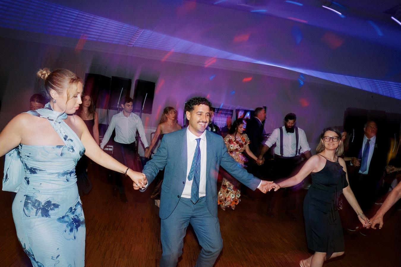 A group of people in formal attire joyfully dancing in a circle at a vibrant indoor event, surrounded by colorful lighting effects.