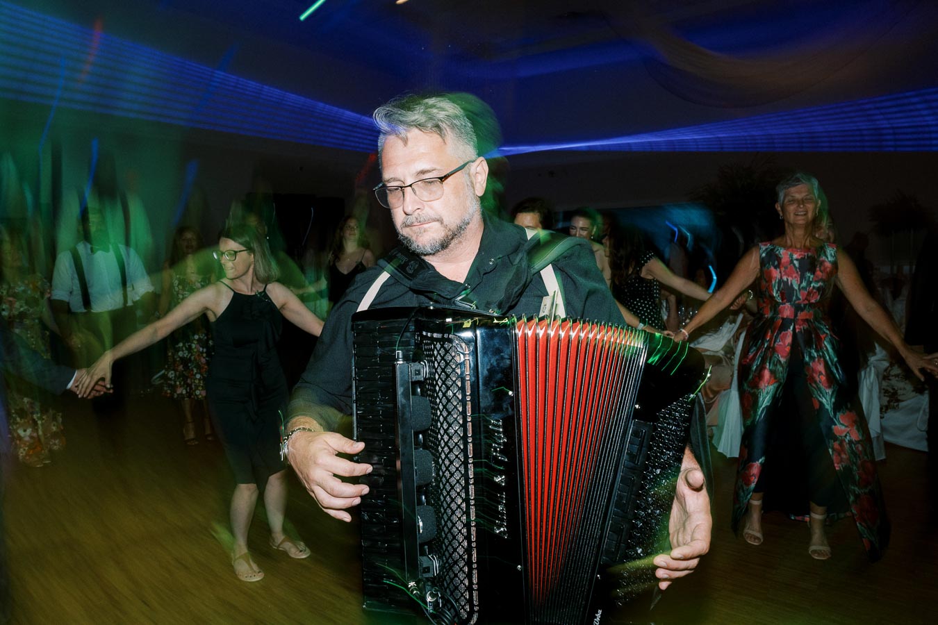 A man playing an accordion at a lively party, surrounded by people dancing in colorful lighting.