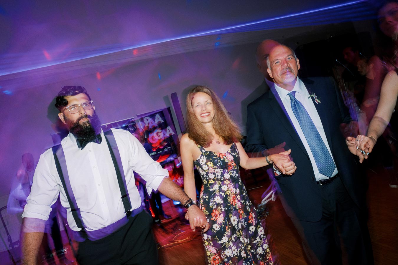 Three people holding hands and dancing at a celebration, with colorful lights creating a lively atmosphere. The man on the left wears suspenders and glasses, the woman in the center is in a floral dress, and the man on the right is in a suit with a boutonniere.