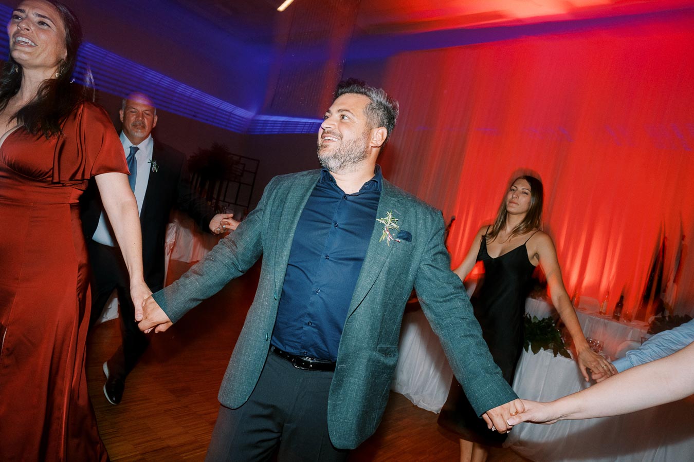 A group of people joyfully dancing in a warmly lit event hall, with vibrant red and blue lighting creating a lively atmosphere.