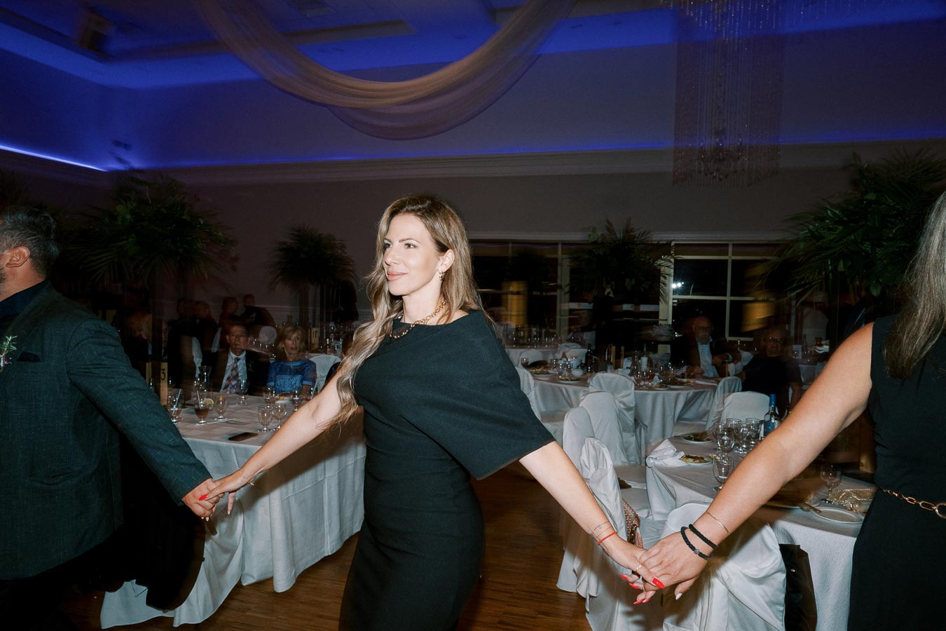 A woman in a black dress holding hands with others, participating in a dance at an elegant indoor event with tables set for dining, ambient lighting, and decorative plants in the background.