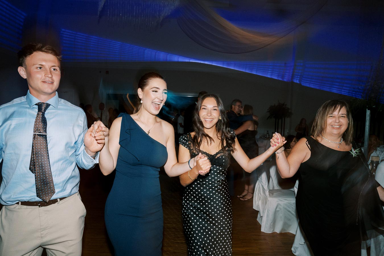 A group of people joyfully dancing at an indoor party, dressed in formal attire and holding hands, with blue ambient lighting and a decorated venue in the background.