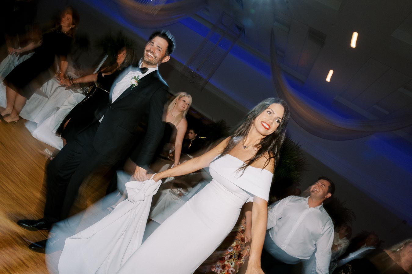 Smiling couple dancing at a wedding reception, with the groom in a black tuxedo and the bride in an off-shoulder white gown, surrounded by guests under soft lighting.