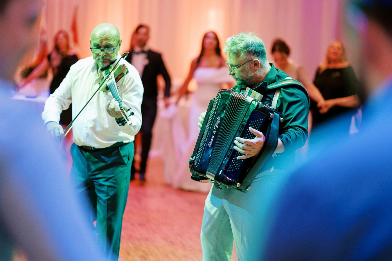 A violinist and accordionist perform lively music at a wedding reception, with guests dancing joyfully in the background.