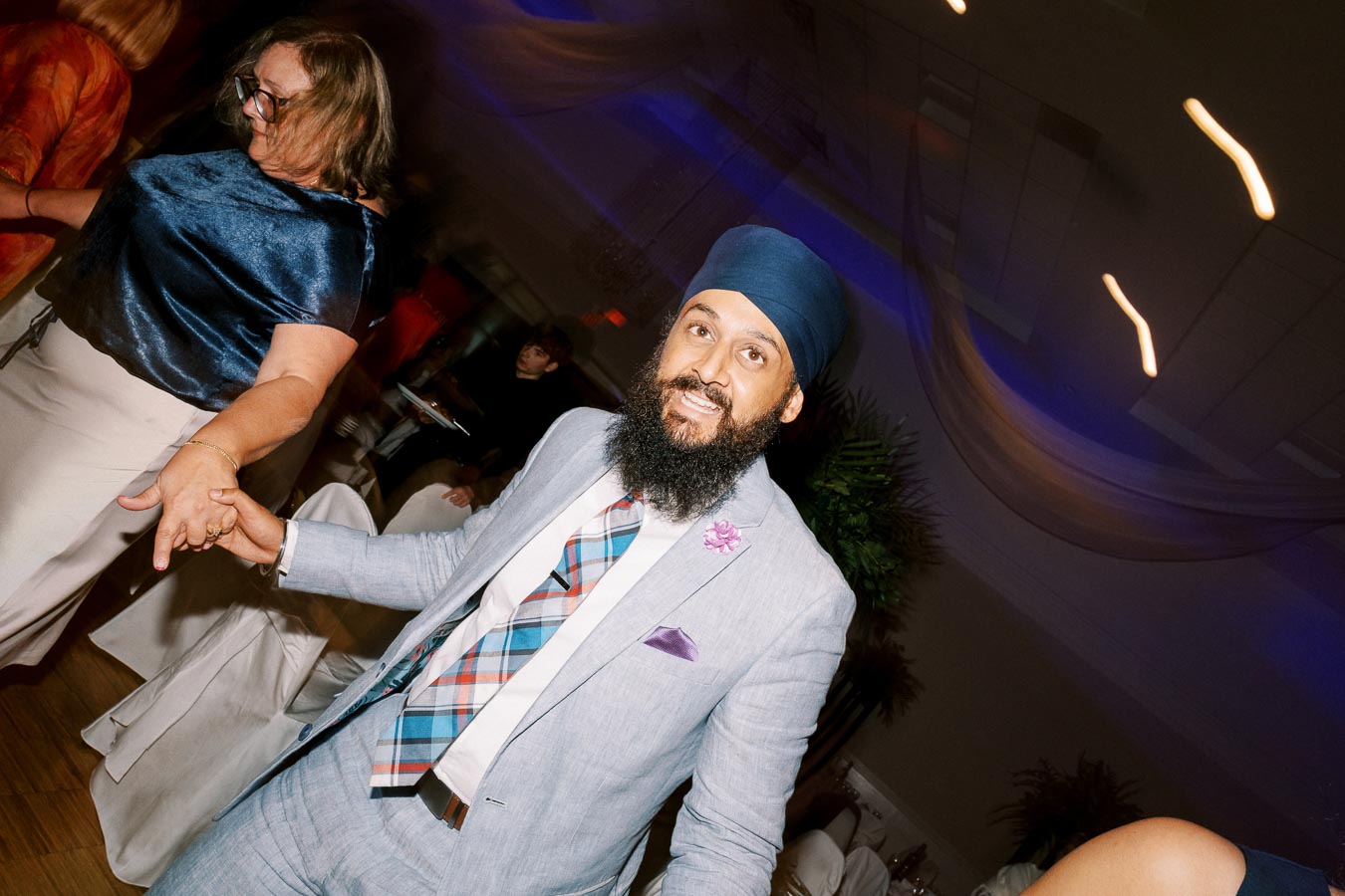 Man in a suit and turban dancing at a lively indoor event, smiling and holding hands with another person, surrounded by other guests in a warmly lit venue.