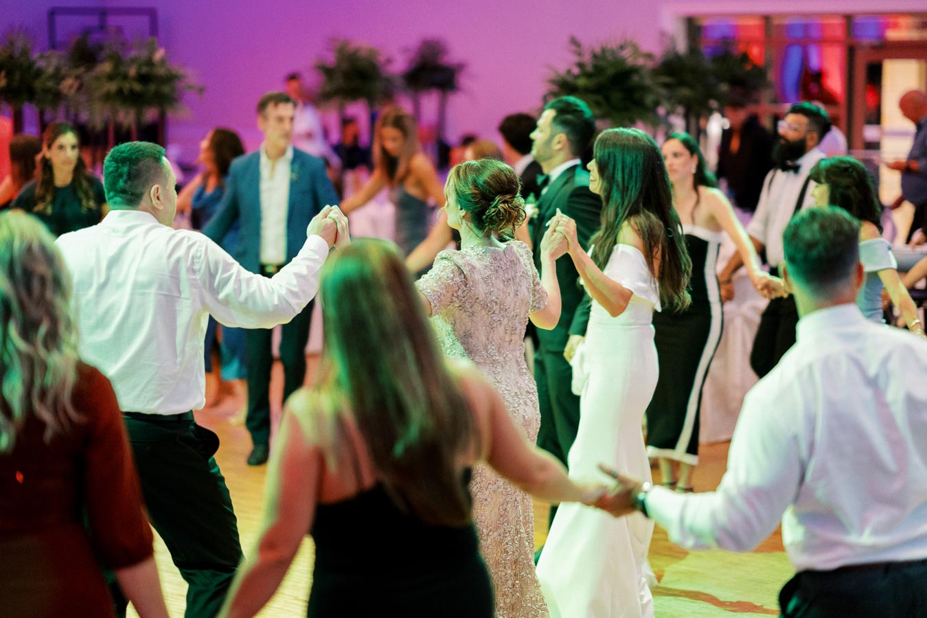 A group of elegantly dressed people dancing in a circle at a festive indoor event, with vibrant lighting and lively atmosphere, suggesting a celebration or wedding reception.