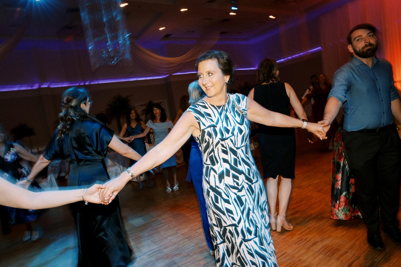 Group of people dancing in a circle at a lively indoor event, featuring a woman in a patterned dress in the center, under colorful lighting.