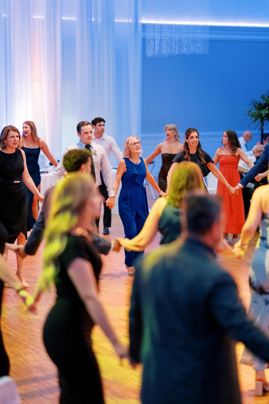 A group of people holding hands and dancing in a circle at a formal event in a ballroom, with blue lighting and elegant attire, creating a joyful and festive atmosphere.