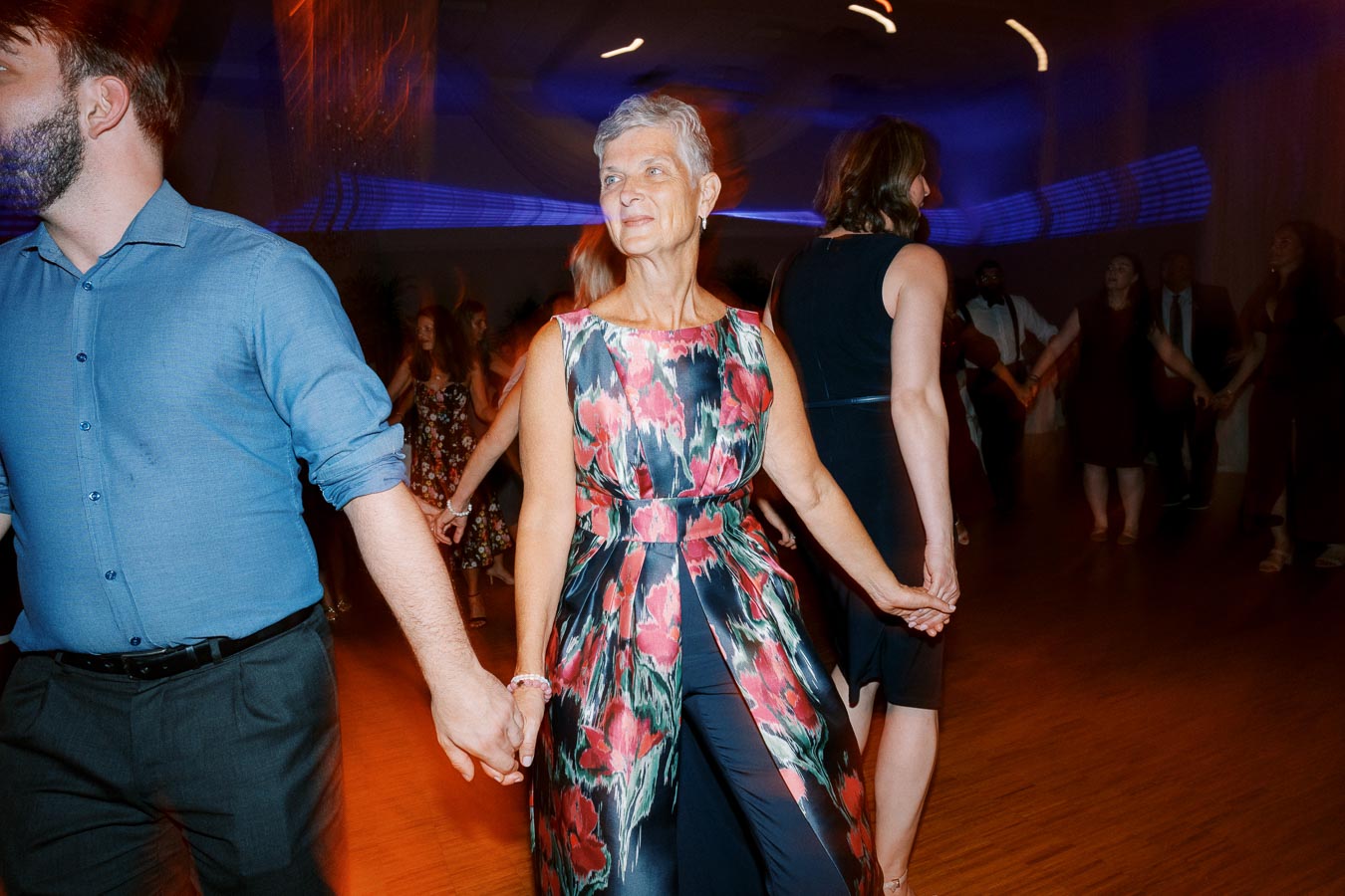 Senior woman in a floral dress dancing joyfully at a social event, holding hands with other people on the dance floor.