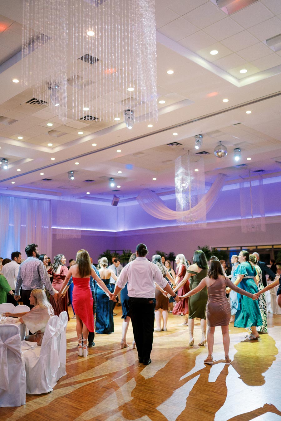 A lively group of people dancing in a decorated event hall under chandeliers with elegant lighting.