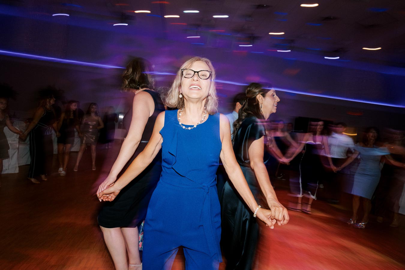 Group of people joyfully dancing in a bright room, with a woman in a blue dress and glasses in the foreground, holding hands with others. The vibrant lighting creates a festive atmosphere.